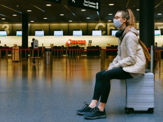 Woman sitting on luggage in airport wearing a mask