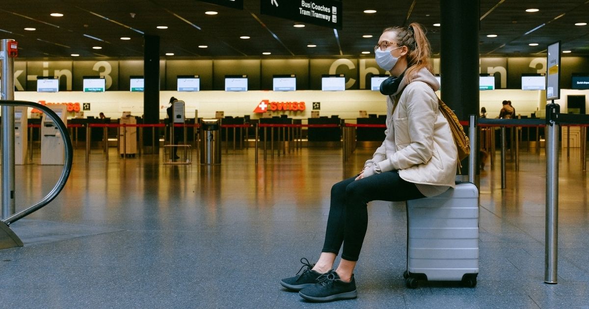 Woman sitting on luggage in airport wearing a mask