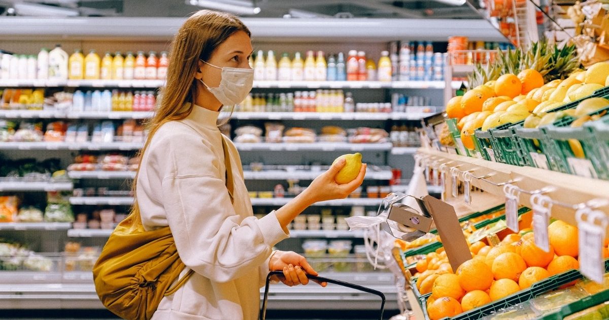 Woman in a yellow tshirt, beige jacket and mask picking out fruit.