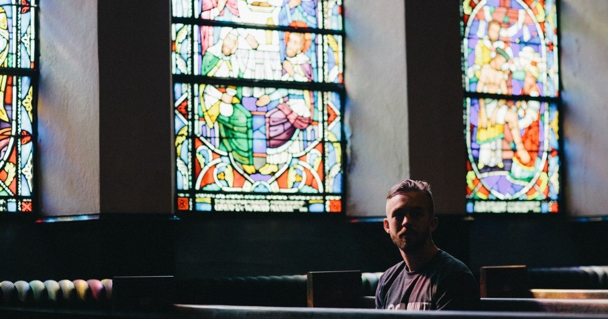 Man sitting in church pew backed by stained glass windows