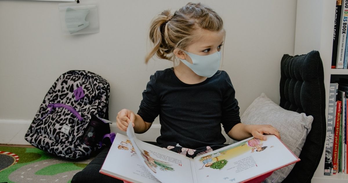 girl with mask on and backpack reading a book