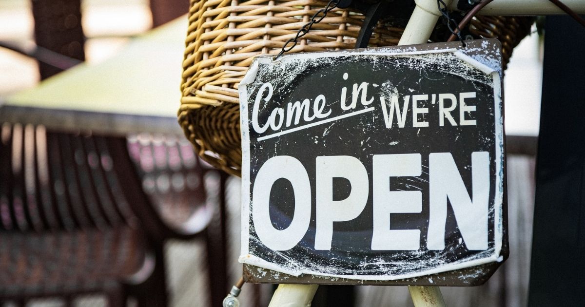 Black and white "Come in We're Open" sign pinned to a white bicycle with a wicker basket.