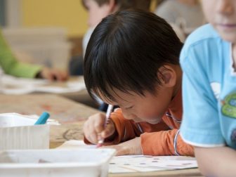 Captured in a metropolitan Atlanta, Georgia primary school, seated amongst his classmates, this photograph depicts a young Asian-American schoolboy, who was in the process of drawing with a pencil on a piece of white paper atop a table that had been covered by a layer of removable brown paper.