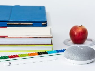 A stack of school books, an iPad Pro in a saturated blue Snugg case, an Apple Pencil, a multicolored Lego ruler, a Google Home Mini processing a command, and a red apple encircled by the Google Home power cord.