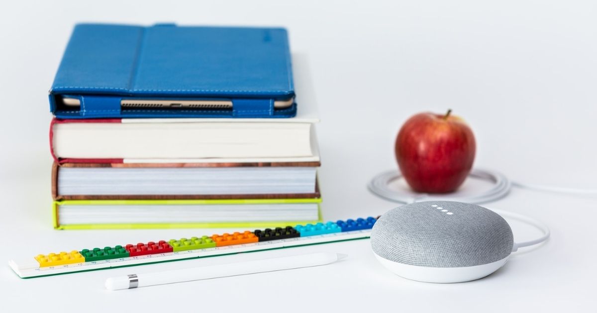 A stack of school books, an iPad Pro in a saturated blue Snugg case, an Apple Pencil, a multicolored Lego ruler, a Google Home Mini processing a command, and a red apple encircled by the Google Home power cord.