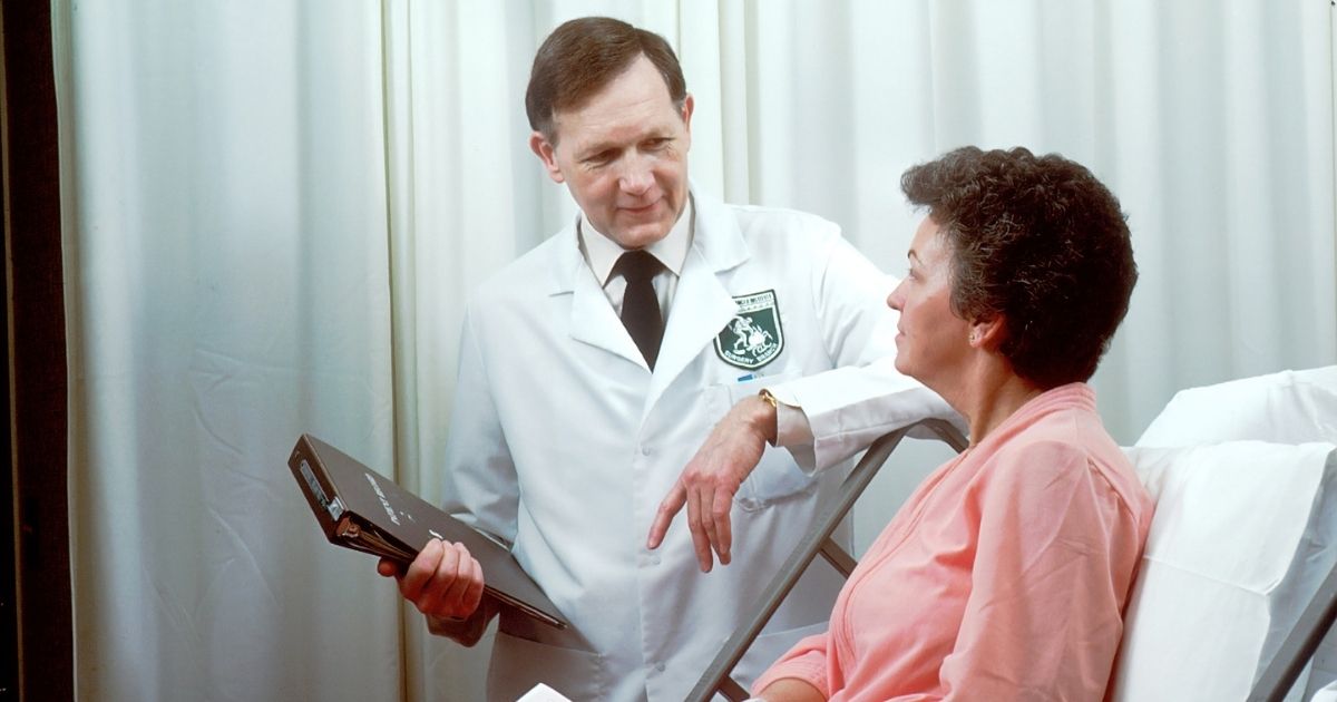 A Caucasian male doctor from the Oncology Branch consults with a Caucasian female adult patient, who is sitting up in a hospital bed.