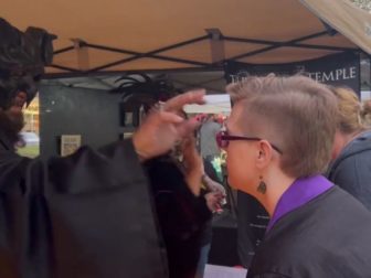 A man dressed in black, flowing robes and a black horned headdress paints an upside-down cross on a festivalgoer's forehead before exchanging the phrase, "Hail, Satan."