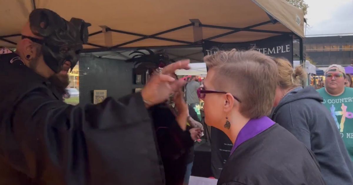 A man dressed in black, flowing robes and a black horned headdress paints an upside-down cross on a festivalgoer's forehead before exchanging the phrase, "Hail, Satan."