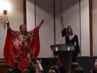 A man in a red caped suit and a woman in black exhort a crowd at a gathering of The Satanic Temple at a Boston hotel.