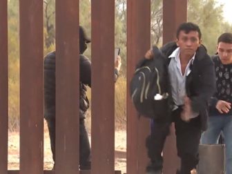Illegal aliens pour through a breach in the border wall in Lukeville, Arizona, while a human smuggler records their faces.