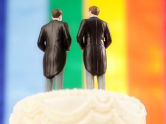 This image shows the top of a traditional wedding cake, with two men in tuxedos and tails as the wedding cake topper and with a rainbow "pride" flag in the background.